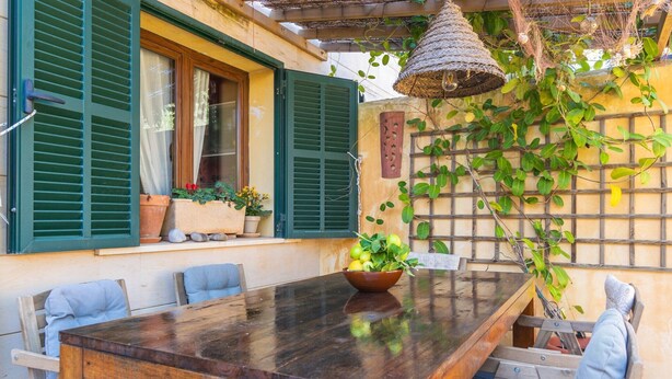 An outdoor dining area with a wooden table and chairs, a potted plant, and a hanging light fixture.