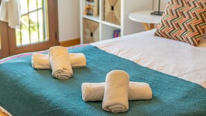 A neatly made bed with a teal blanket and rolled white towels, a white bedside table with a lamp, and a colorful chevron pillow.