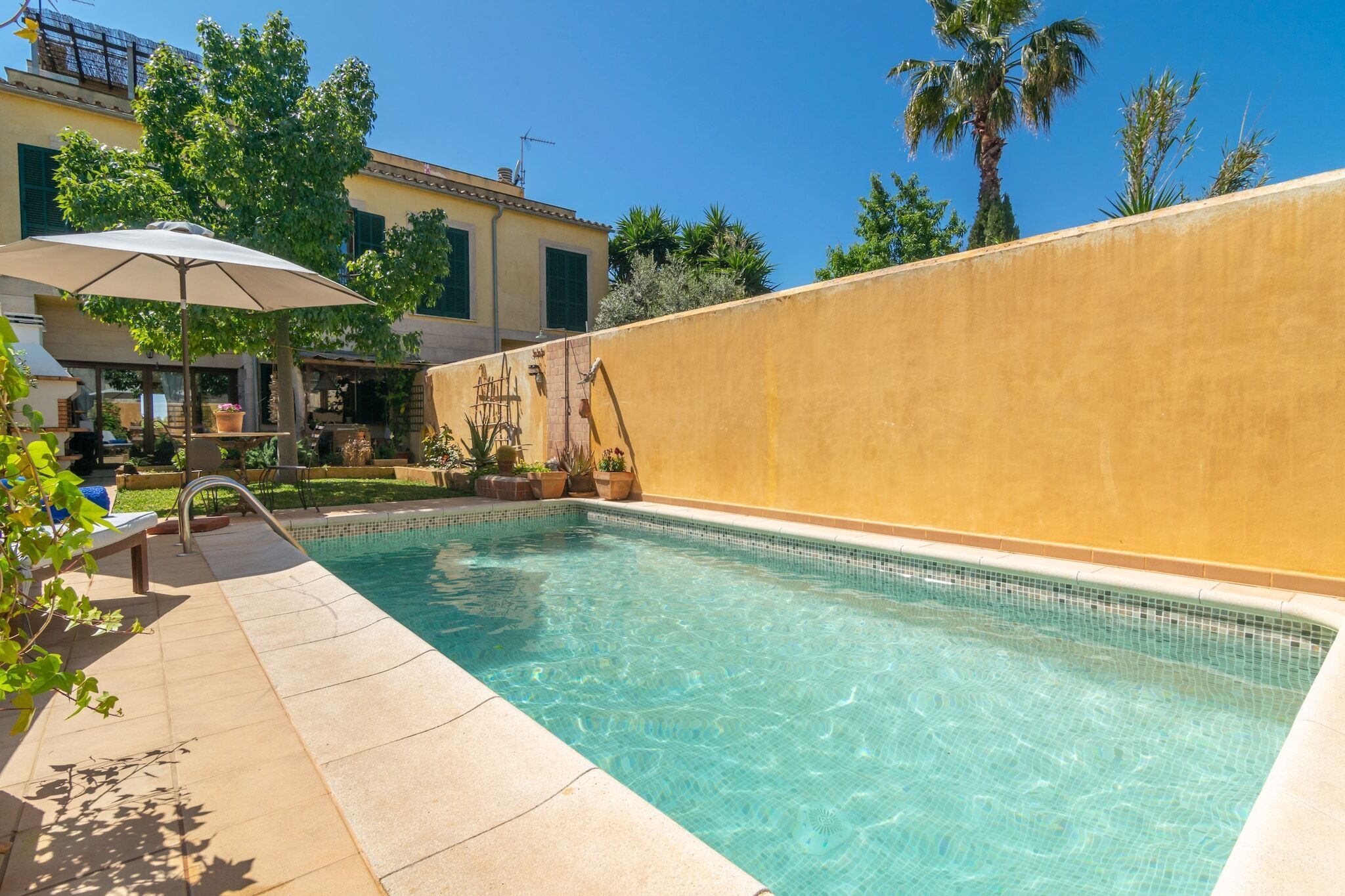 A swimming pool with clear water, surrounded by a yellow wall and a patio area with a table and chairs.