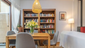 A cozy room with a wooden dining table, grey chairs, a bookshelf, a potted plant with flowers, a hanging light fixture, and a bed with a white cover.