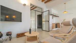 A modern living room with a round wooden table, two cushioned chairs, a large flat-screen TV, and a kitchen visible through an open door.