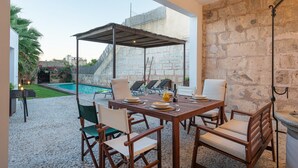 An outdoor dining area with a stone wall backdrop, a covered seating area, and a pool visible in the background.