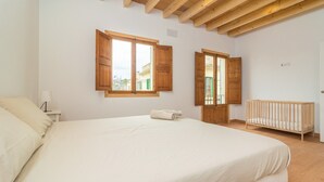 A bedroom with a large bed, wooden ceiling beams, two windows with wooden shutters, a crib, and a small bedside table with a lamp.