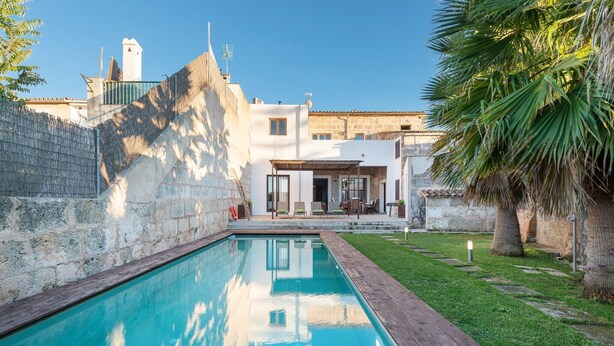 A swimming pool with a wooden deck, a stone wall, and a building with a patio area in the background.