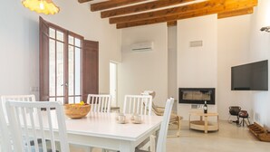 A dining area with a white table and chairs, a wooden bowl with fruits, a television, and a fireplace.