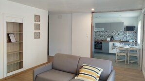 A modern living room with a grey sofa, a wooden bookshelf, and a kitchen in the background.