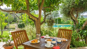 An outdoor wooden table with chairs, a potted plant, and a breakfast setting with croissants, coffee, and a teapot.