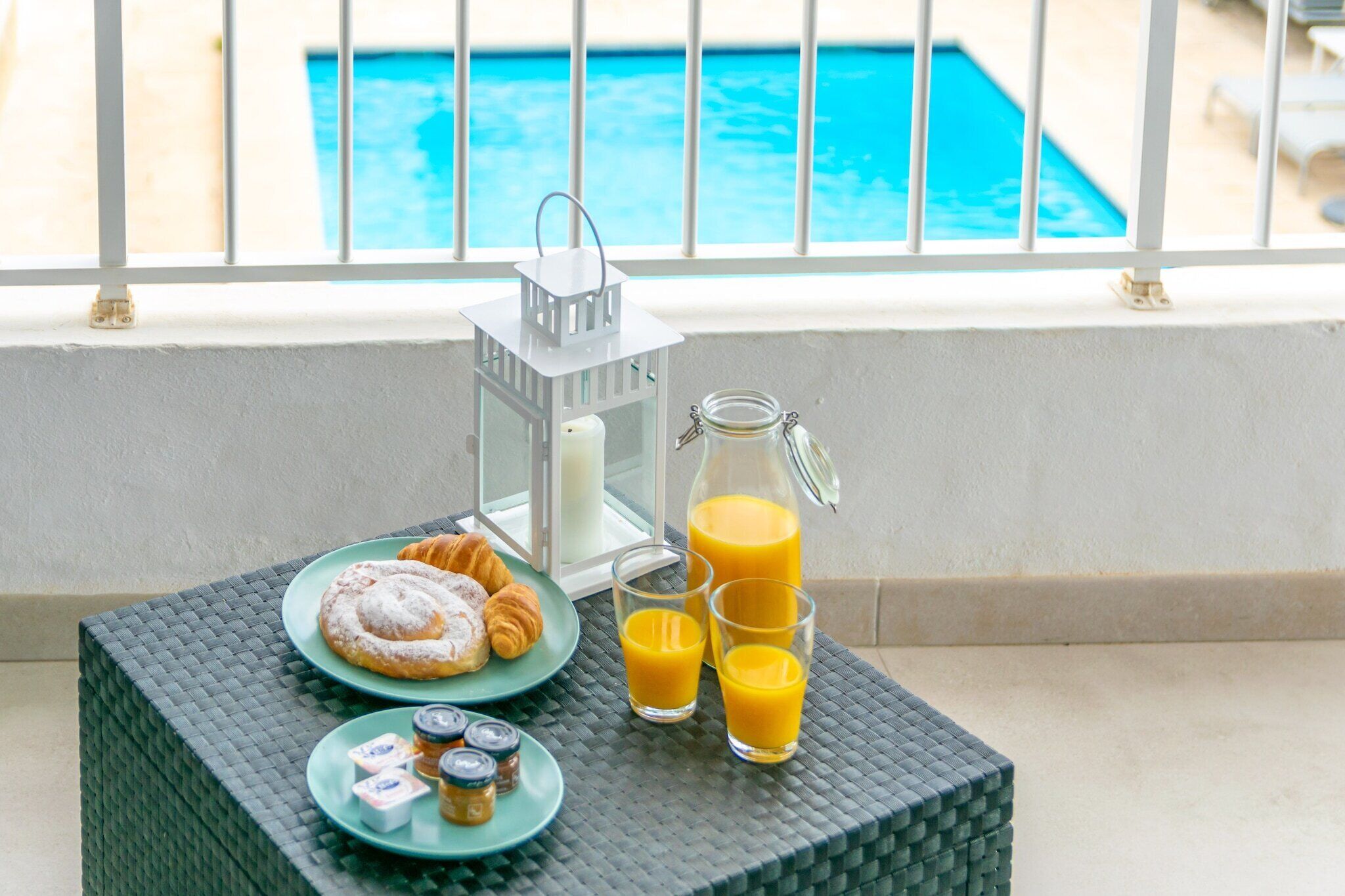 Morgenmadsbord med croissanter, kager og juice på en balkon med udsigt til poolen.