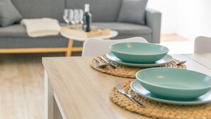A dining table with two teal plates and cutlery, set on a woven placemat, in a modern living room with a grey sofa and a small round table with wine glasses and a bottle. - Ona 2 - Apartment In Cala D'or. Free Wifi (, Illes Balears)