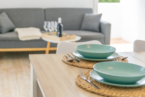 A dining table with two teal plates and cutlery, set on a woven placemat, in a modern living room with a grey sofa and a small round table with wine glasses and a bottle.