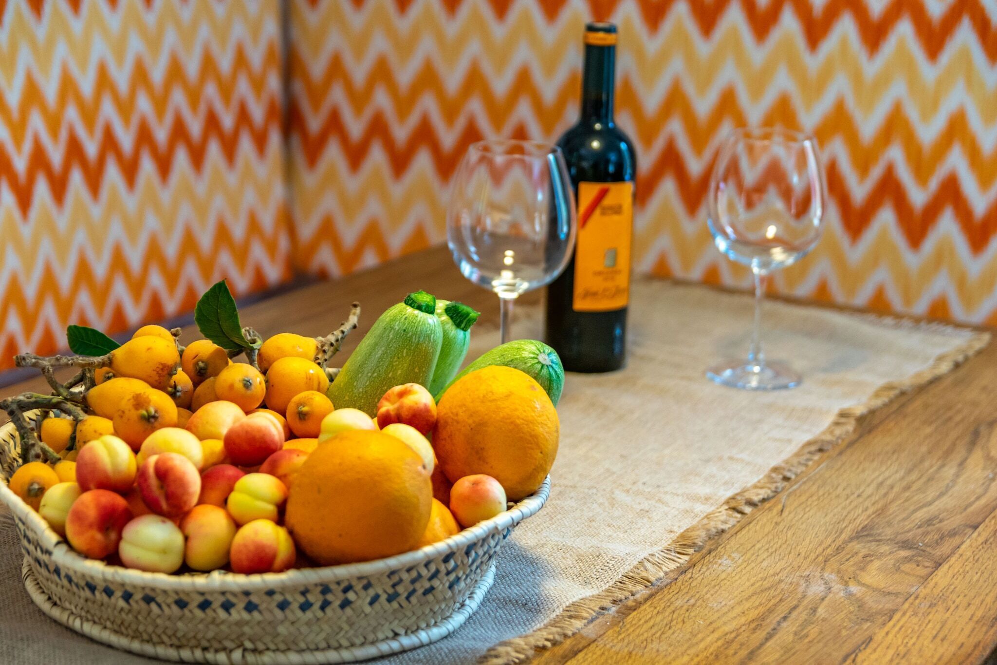 A table with a fruit basket containing peaches, oranges, and a zucchini, a bottle of wine, and two empty wine glasses.