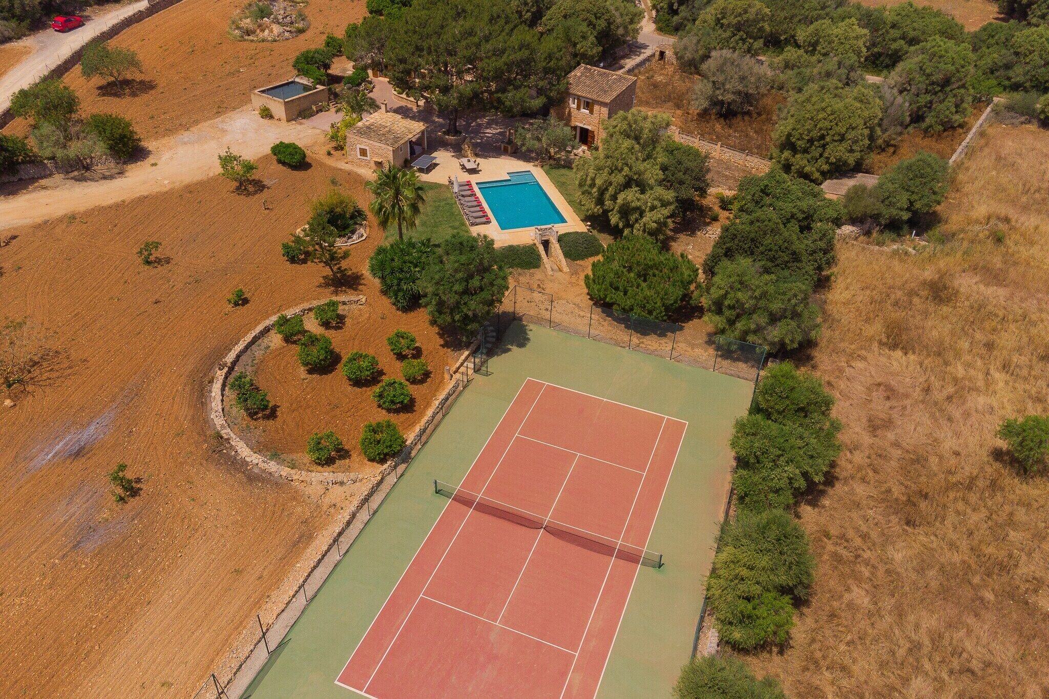 Vue aérienne d’une propriété comprenant un court de tennis, une piscine et plusieurs bâtiments entourés d’arbres.