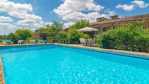 Ein klares, blaues Schwimmbecken mit Liegestühlen und einem Tisch mit Sonnenschirm, umgeben von viel Grün und traditioneller Architektur.