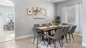 Un coin repas avec une table rectangulaire, six chaises et un lustre. Des œuvres d’art encadrées ornent le mur, et une fenêtre est équipée de stores.