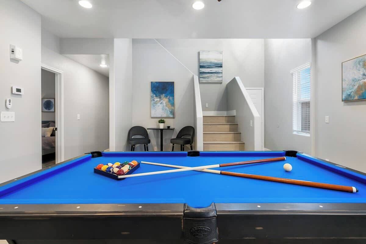 Une table de billard avec des billes et des queues, un escalier, et deux chaises dans une pièce aux murs blancs et à la table de billard bleue.