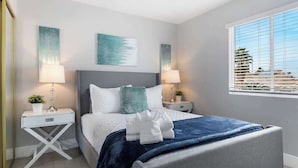 Une chambre à coucher comprenant un lit, deux tables de chevet, une commode, une fenêtre avec des stores et des œuvres d’art accrochées aux murs.