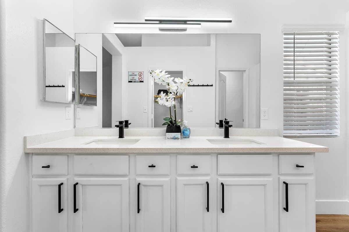 A modern bathroom with white cabinetry, two sinks, a large mirror, and a potted orchid.