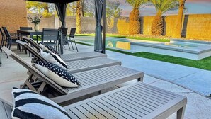 Un espace extérieur aménagé, abrité et doté de fauteuils confortables et d’une table, ainsi qu’une piscine et des palmiers en arrière-plan.