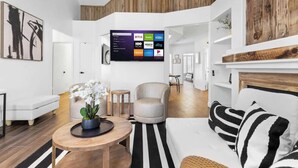 Un salon moderne doté d’un téléviseur à écran plat diffusant des services de streaming, d’une table basse ronde en bois sur laquelle est posée une orchidée en pot, d’un fauteuil gris et d’un coussin rayé sur un canapé.