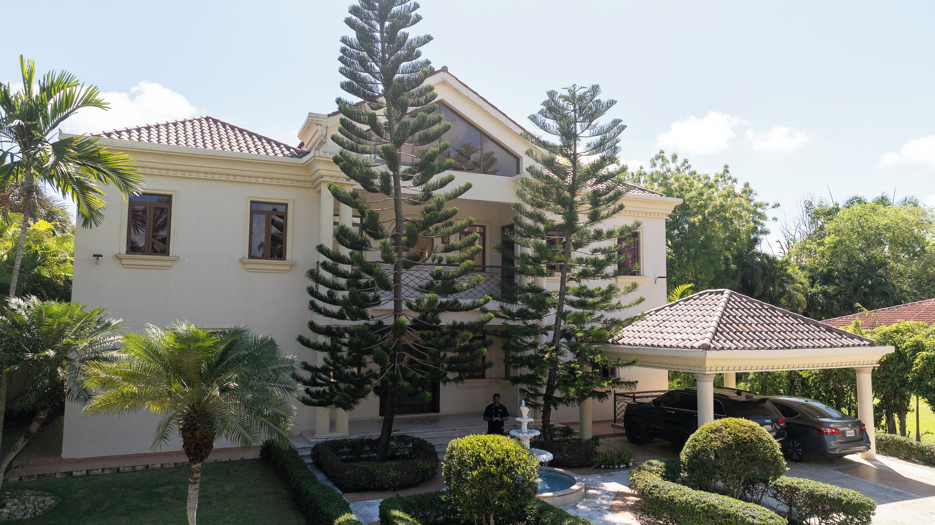 A two-story house with a tiled roof, a covered porch, and a well-maintained garden with palm trees and a fountain.