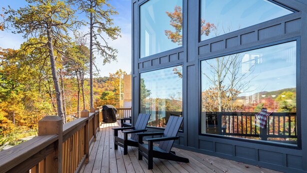 A wooden deck with Adirondack chairs, large windows, and a view of autumn trees.