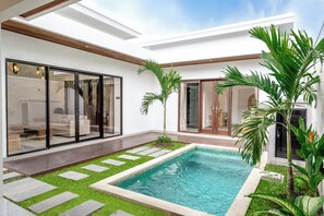 A modern outdoor pool area with a glass-enclosed patio, a small garden with palm trees, and a clear blue pool.