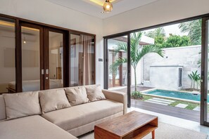 Ein modernes Wohnzimmer mit einem beigefarbenen Sofa, einem Holztisch und einer Glastür, die zu einem Poolbereich mit Steinwand und viel Grün führt.