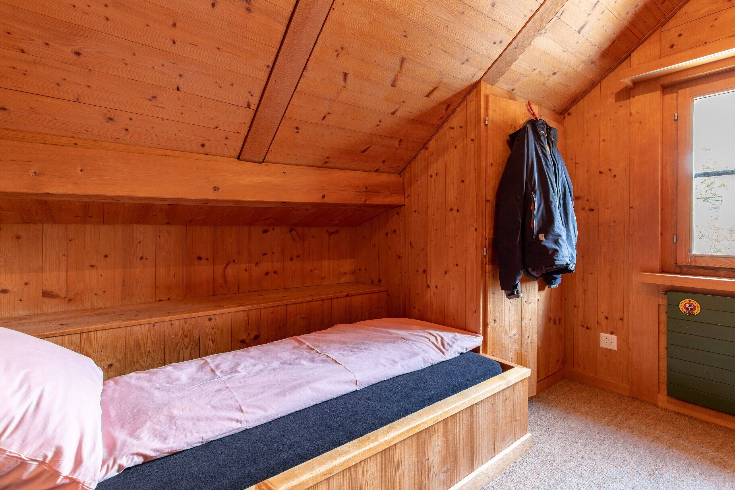 A small, wooden cabin room with a single bed, a bench, and a coat rack.