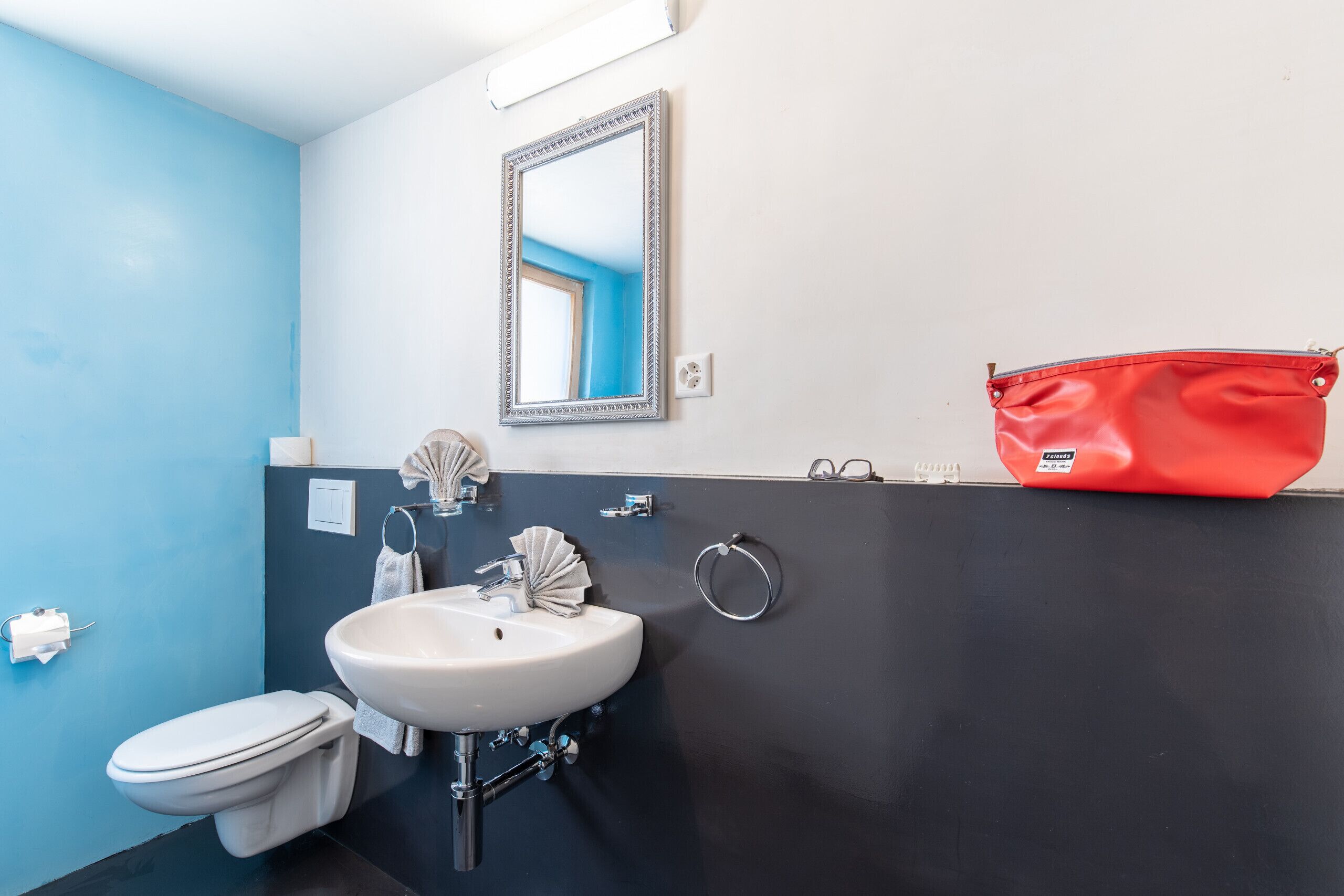 Une salle de bain avec un lavabo blanc, des toilettes, un miroir et un sac rouge posé sur le comptoir.