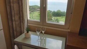 A small dining area with a table, two wine glasses, and a potted plant. A window offers a view of a green landscape and the sea.