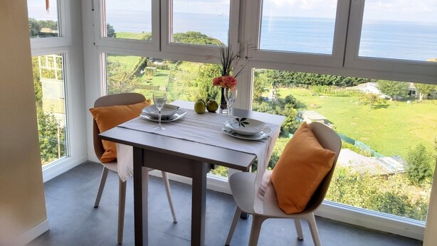 A dining area with a table set for two, overlooking a scenic view of a green landscape and the sea.