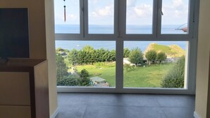 A room with a large window overlooking a coastal landscape, including a grassy area and a view of the sea.