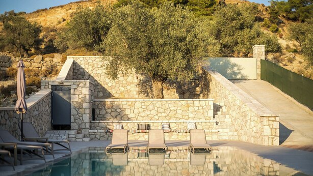 Piscina de piedra con sillas de descanso, pared de piedra y vistas a olivos y colinas.