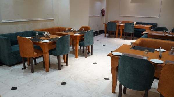 A dining area with wooden tables and teal chairs, a fire extinguisher on the wall, and a white tiled floor.