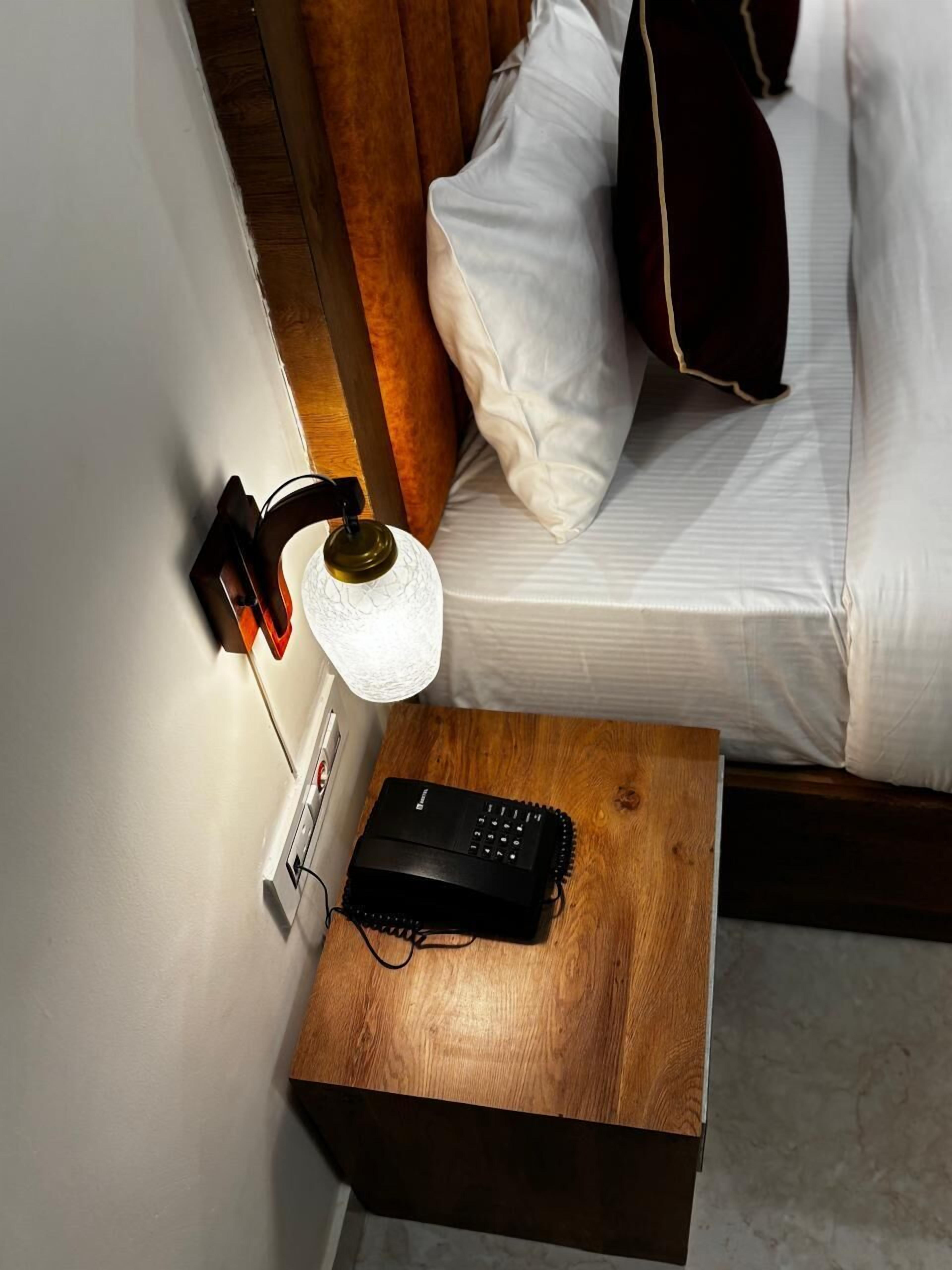 Un lit bien fait, avec du linge de lit blanc et un oreiller marron foncé, une table de chevet avec une lampe et une télécommande, et une tête de lit en bois.