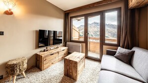 Ein Zimmer mit großem Fenster, einem Flachbildfernseher, einem Holztisch, einer Couch und einem kleinen Hocker.