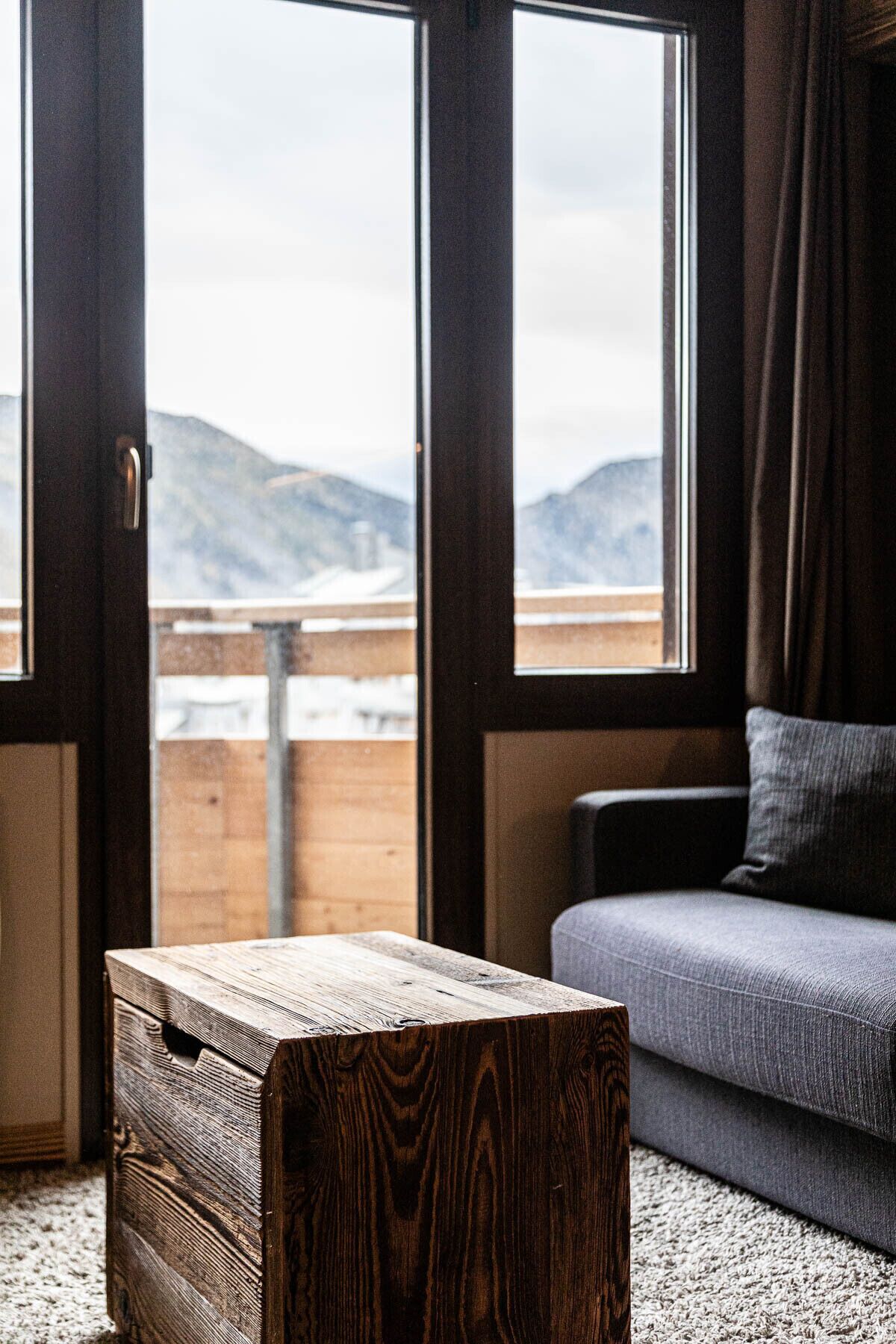 A room with a wooden chest, a grey sofa, and a view of mountains through large windows.