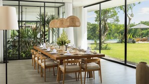 Un coin repas avec une longue table en bois, dressée pour six personnes, entourée de chaises, et une grande fenêtre donnant sur un paysage verdoyant et une piscine.