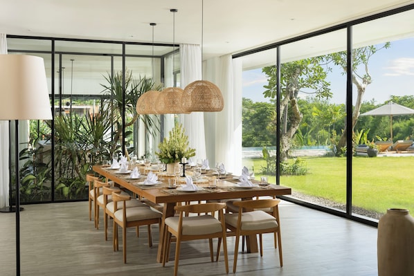 Un coin repas avec une longue table en bois, dressée pour six personnes, entourée de chaises, et une grande fenêtre donnant sur un paysage verdoyant et une piscine.