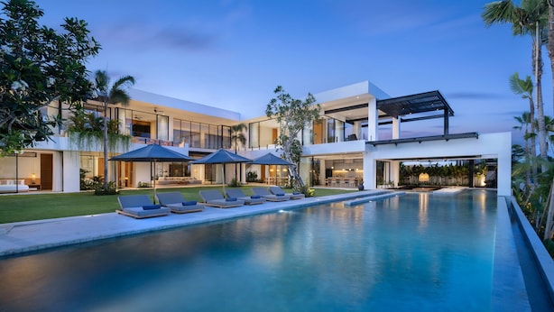 Un espace moderne aménagé au bord de la piscine, avec des chaises longues et des parasols, une grande piscine et un bâtiment contemporain doté de grandes fenêtres et d’un espace salon extérieur.