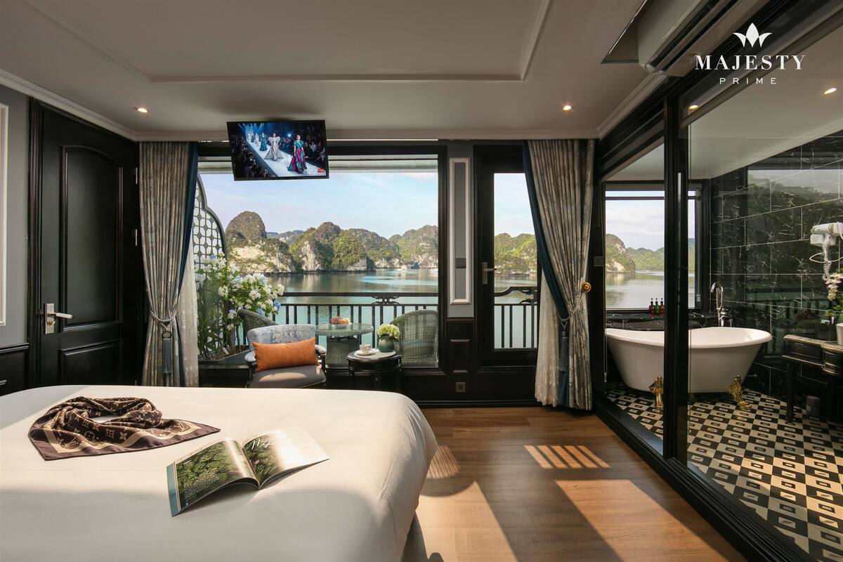 A luxurious hotel room with a large bed, a television, a balcony with a view of the water, and a modern bathroom.