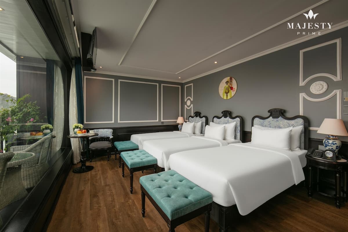 A hotel room with three beds, a small table with a lamp, a window with a view, and a decorative wall piece.