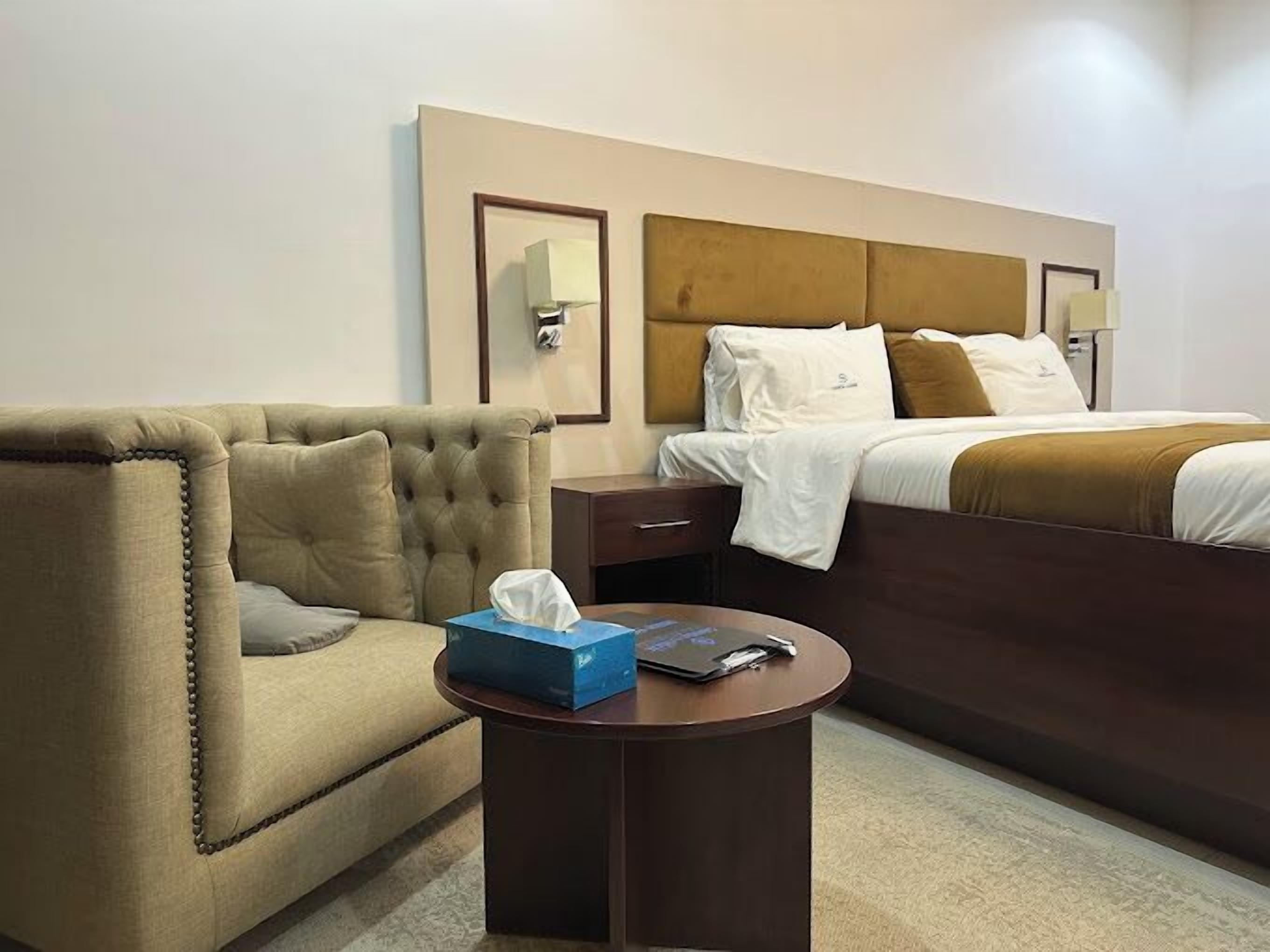 Quarto de hotel com uma cama grande, uma poltrona estofada, uma mesinha de cabeceira com caixa de lenços e controle remoto, e uma luminária fixada na parede.