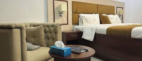 Quarto de hotel com uma cama grande, uma poltrona estofada, uma mesinha de cabeceira com caixa de lenços e controle remoto, e uma luminária fixada na parede.