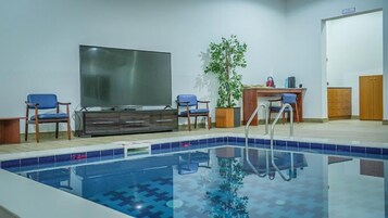 Uma piscina com deck ladrilhado, uma TV de tela plana na parede, duas cadeiras azuis, uma escrivaninha de madeira com planta em vaso e um guarda-roupa de madeira.