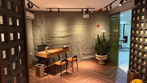 A modern room with a wooden floor, a stone accent wall, a wooden table with chairs, a potted plant, and a yellow fire extinguisher. - Kostkan House (Jakarta)