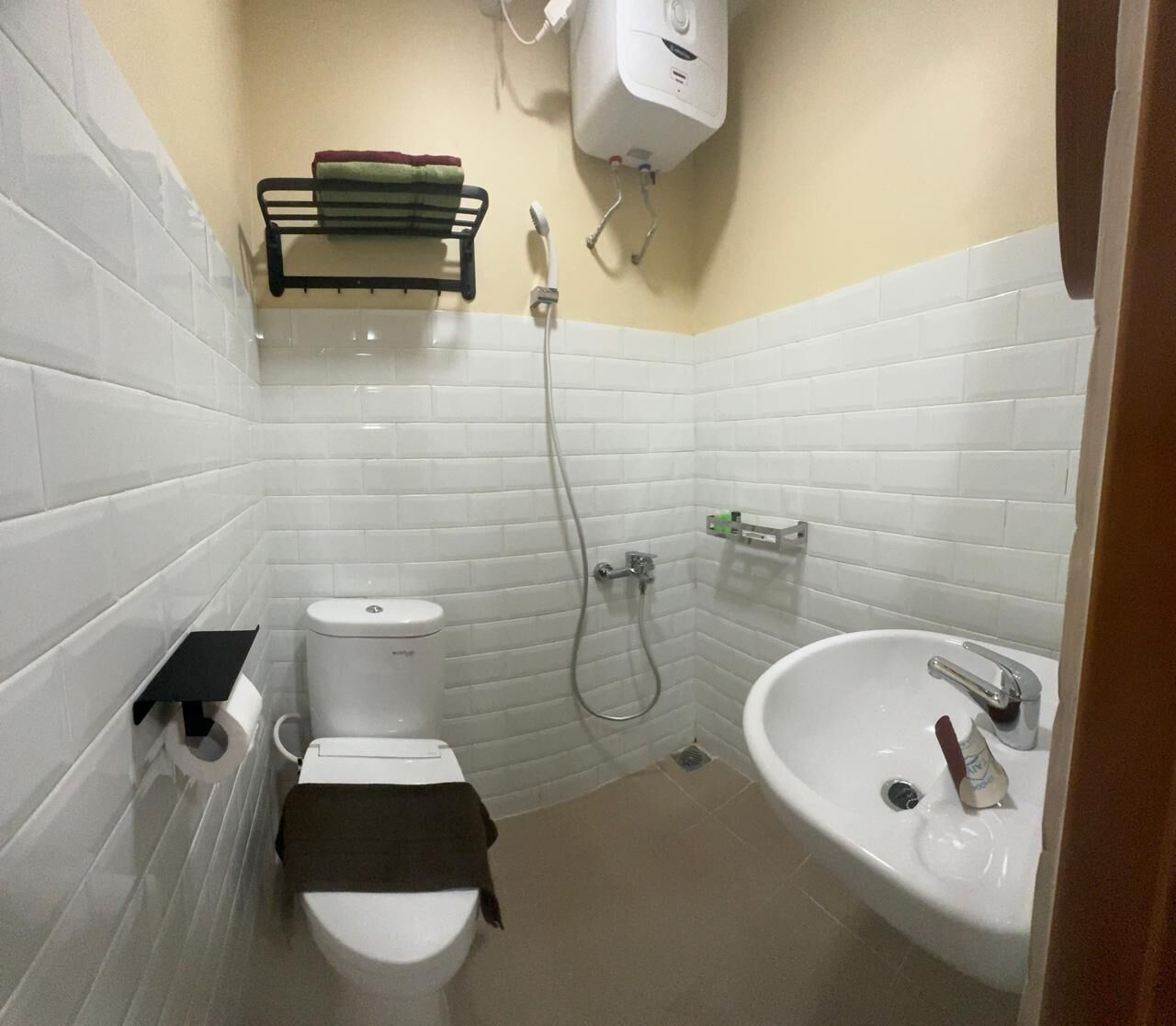 A small bathroom with a toilet, a sink, and a shower. There is a towel rack above the toilet and a soap dispenser on the sink.