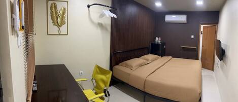 A compact bedroom with a bed, a desk, a chair, a wall-mounted air conditioner, and a door.