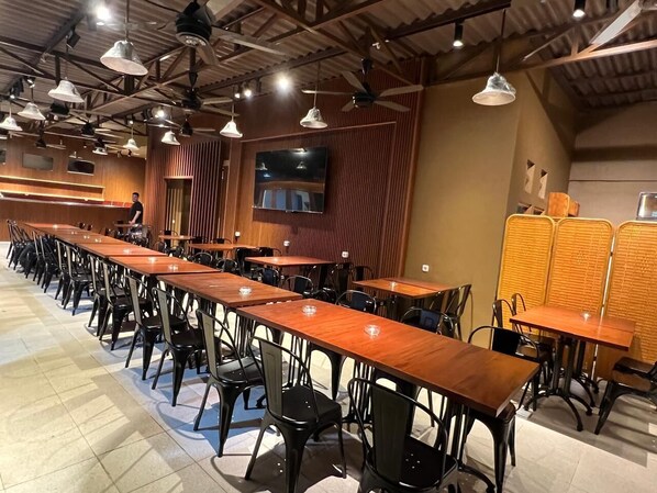 Une grande salle à manger avec des tables en bois et des chaises noires, un écran de télévision et des ventilateurs de plafond.