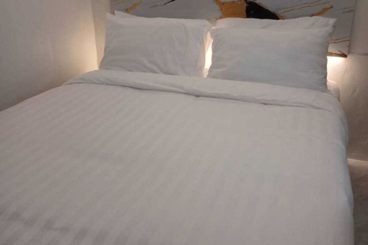 A neatly made bed with white linens and pillows, a headboard with a decorative pattern, and a light-colored wall.